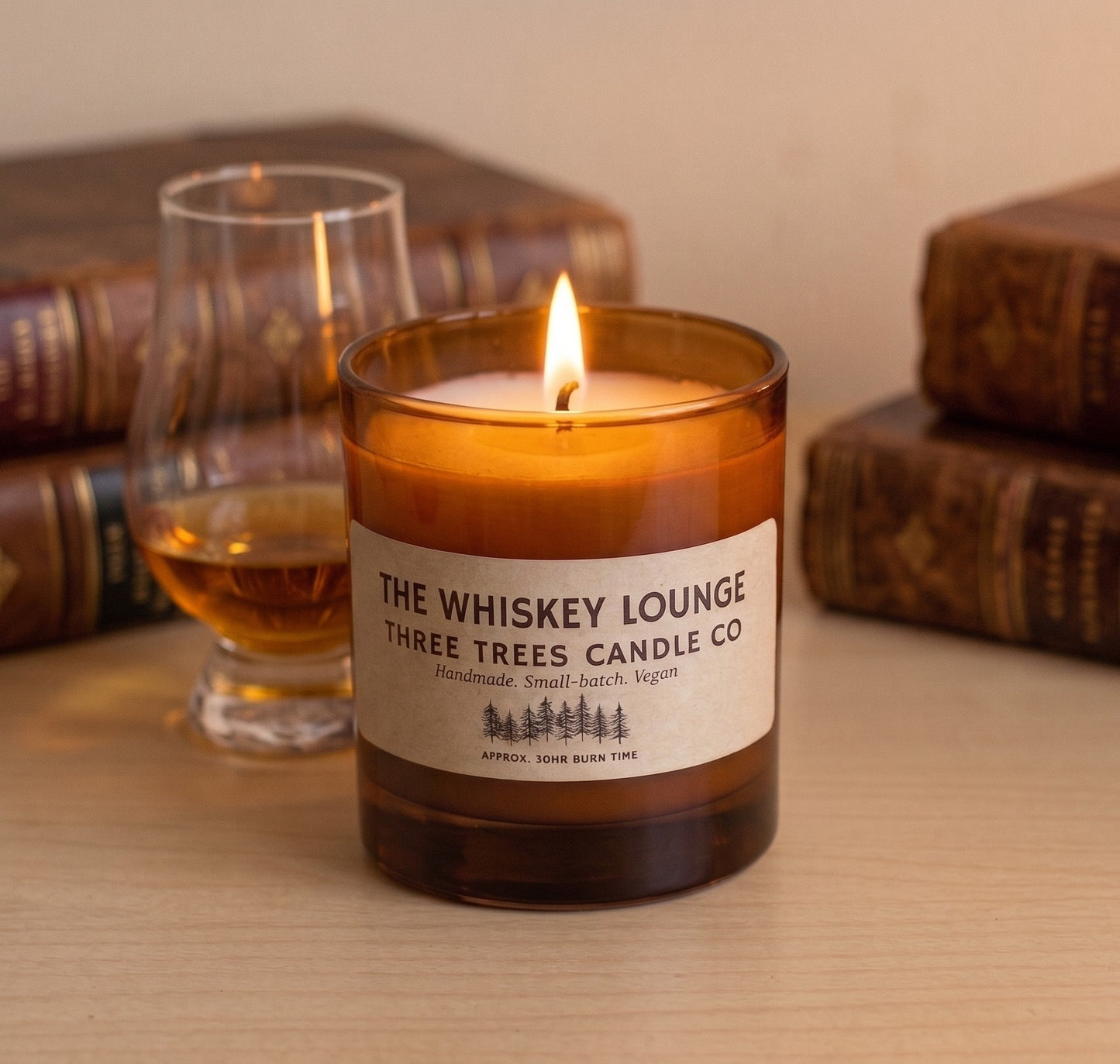 A lit amber glass candle jar labeled 'The Whiskey Lounge' by Three Trees Candle Co, set on a wooden surface with a whiskey glass and vintage books blurred in the background.