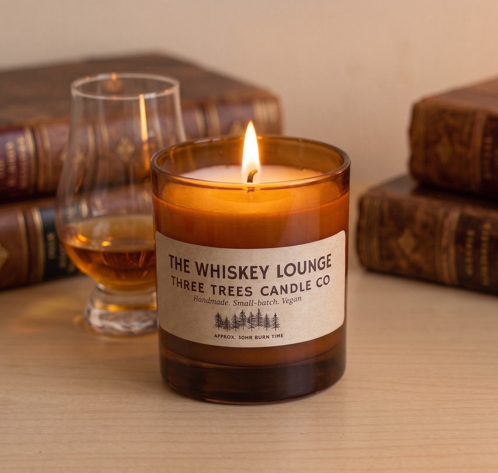 A lit amber glass candle jar labeled 'The Whiskey Lounge' by Three Trees Candle Co, set on a wooden surface with a whiskey glass and vintage books blurred in the background.