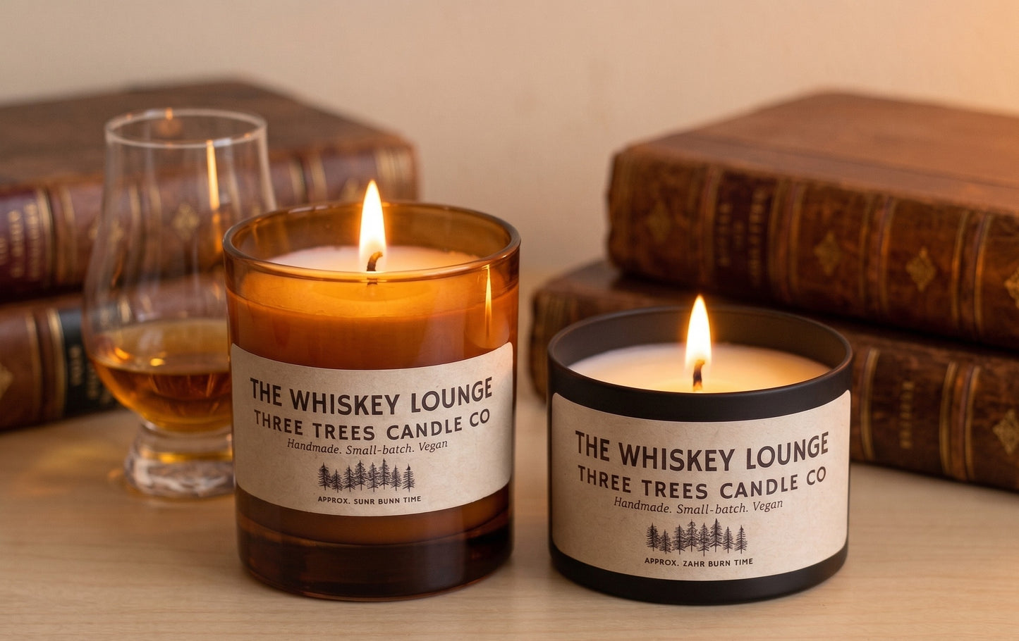 Two lit candles from Three Trees Candle Co. labeled 'The Whiskey Lounge' are displayed on a wooden surface with vintage books and a whiskey glass in the background.